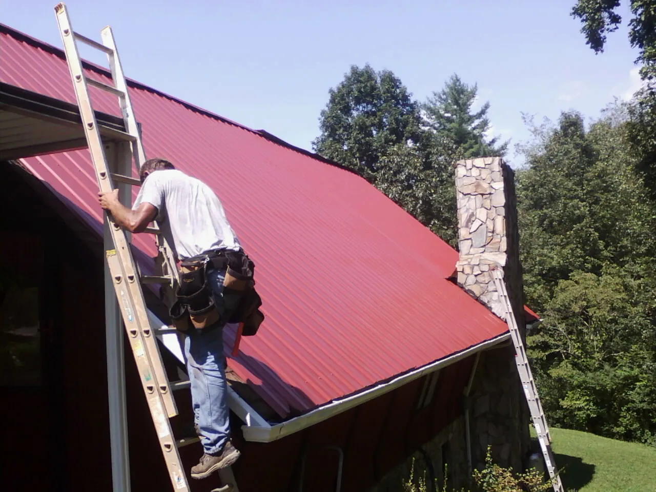 Roofer on a ladder beside a red metal roof and stone chimney — Soffit & Fascia Repair by Roofing Brothers in West Hollywood
